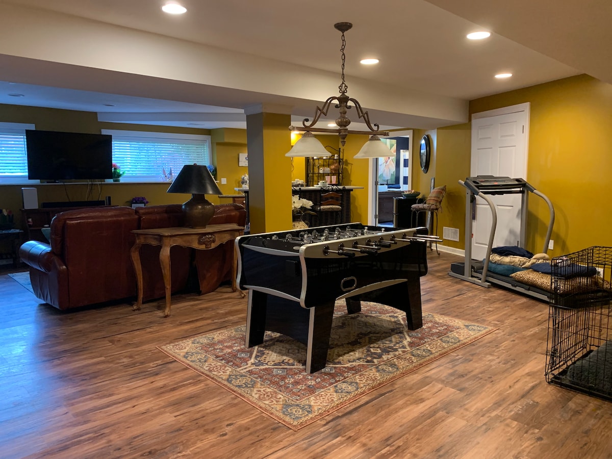 An open living area is showcased, featuring a foosball table at the center. A treadmill is positioned nearby, while two leather sofas are arranged against the wall. Warm yellow walls are complemented by soft overhead lighting, creating a welcoming environment. Large windows allow natural light to fill the space.