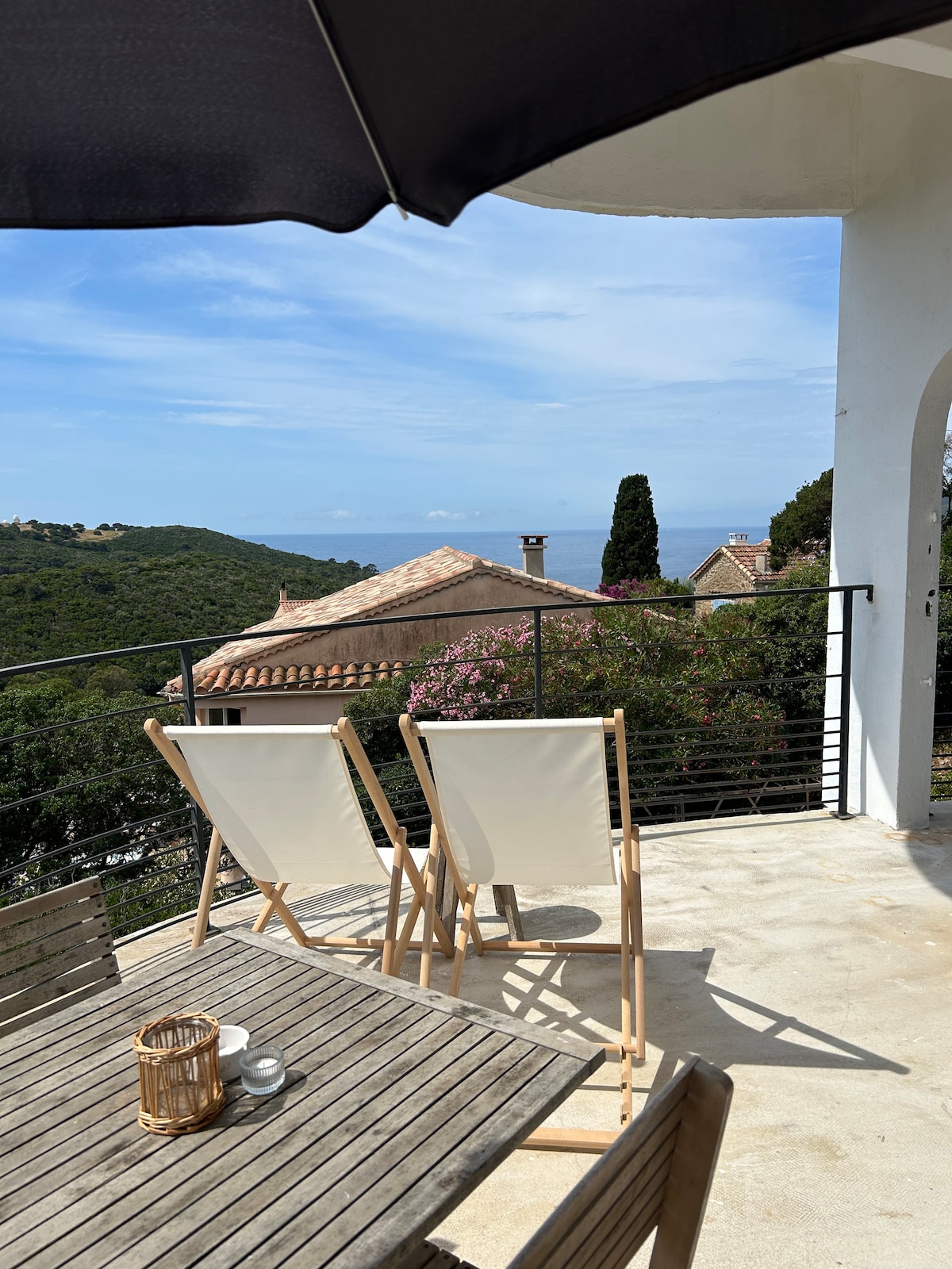 A terrace offers a scenic view overlooking hills and the sea. Two lounge chairs are positioned next to a wooden table, creating a relaxing outdoor seating area. A parasol provides shade, while vibrant greenery and flowers enhance the natural setting.