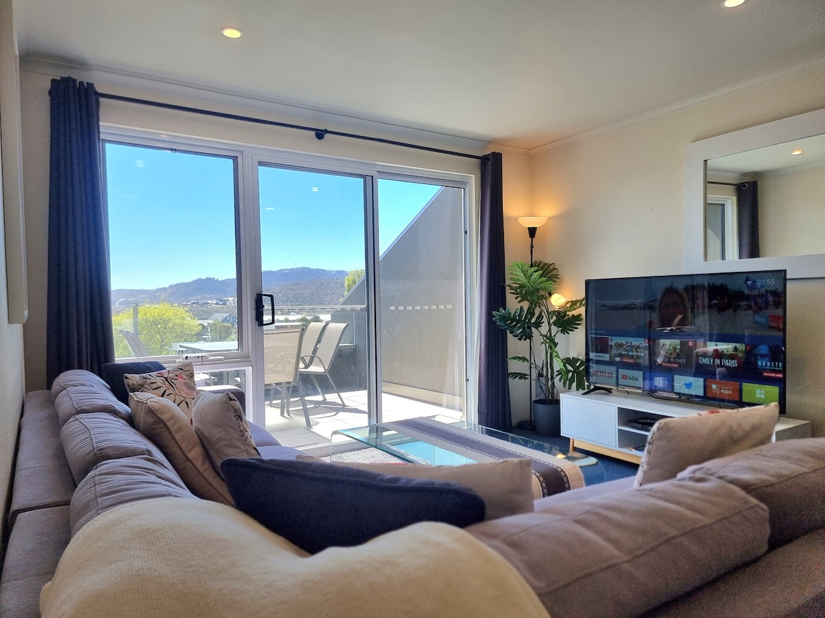 A cozy living area is shown, featuring a comfortable sectional sofa with soft cushions. A large TV sits upon a stylish console, while glass doors lead to a balcony with outdoor seating. Bright natural light fills the space, enhancing the scenic mountain view.