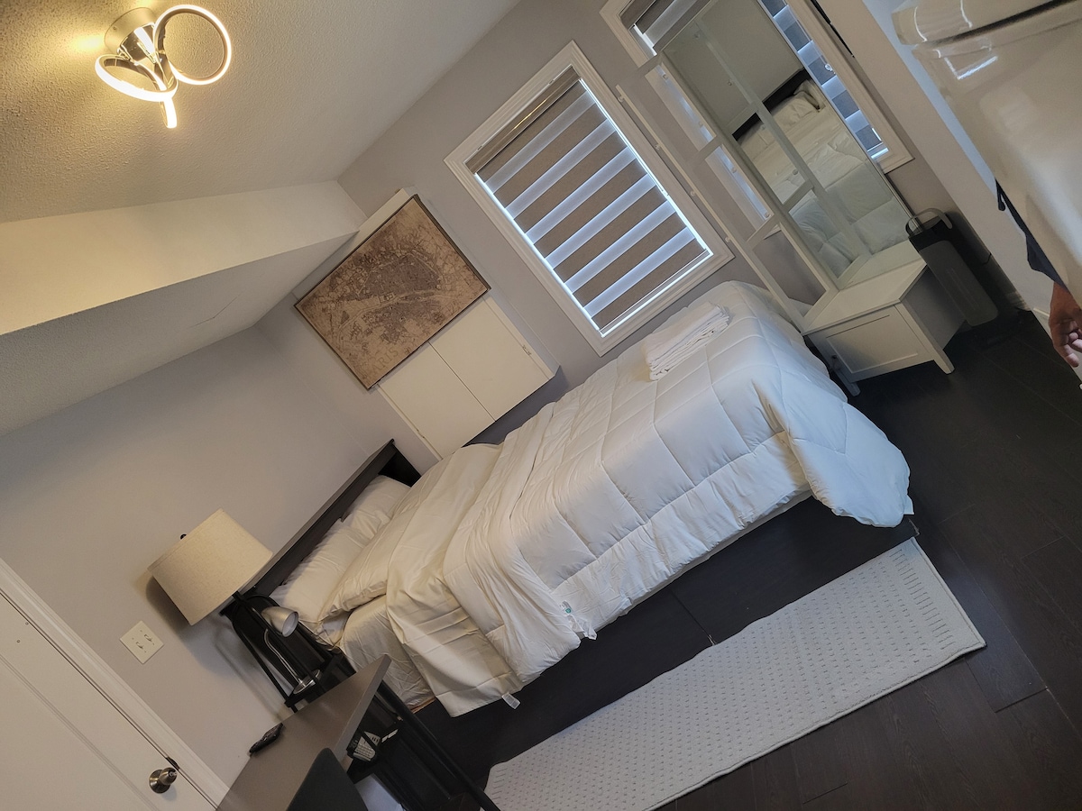 A well-appointed studio room features a queen bed with white bedding and a light-colored lamp on the nightstand. A study desk is situated near the wall across from the bed, and natural light enters through windows with shades. A small area rug adds comfort to the space.