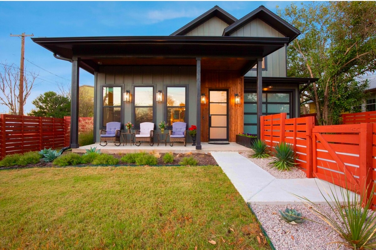 The modern home's exterior is characterized by a striking dark facade and large windows. A covered porch with two chairs invites relaxation, while a well-maintained lawn and vibrant landscaping complement the inviting front entrance. The secure, red fence adds privacy to the property.