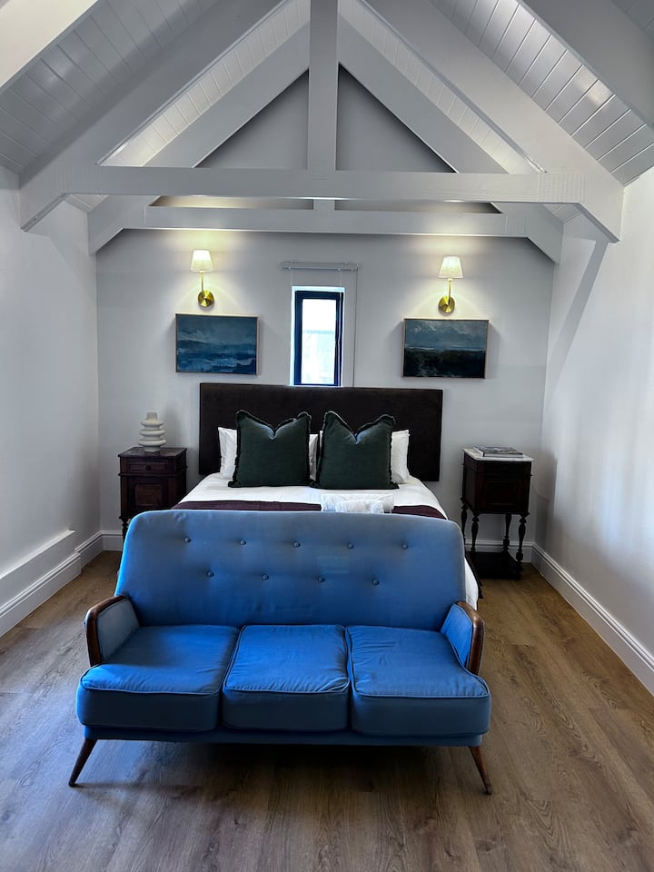 Chambre principale nouvellement construite avec un lit queen size, de hauts plafonds et une lumière naturelle abondante. Comprend un coin salon confortable et des meubles en bois classiques, créant une atmosphère spacieuse et tranquille idéale pour le repos et la détente.