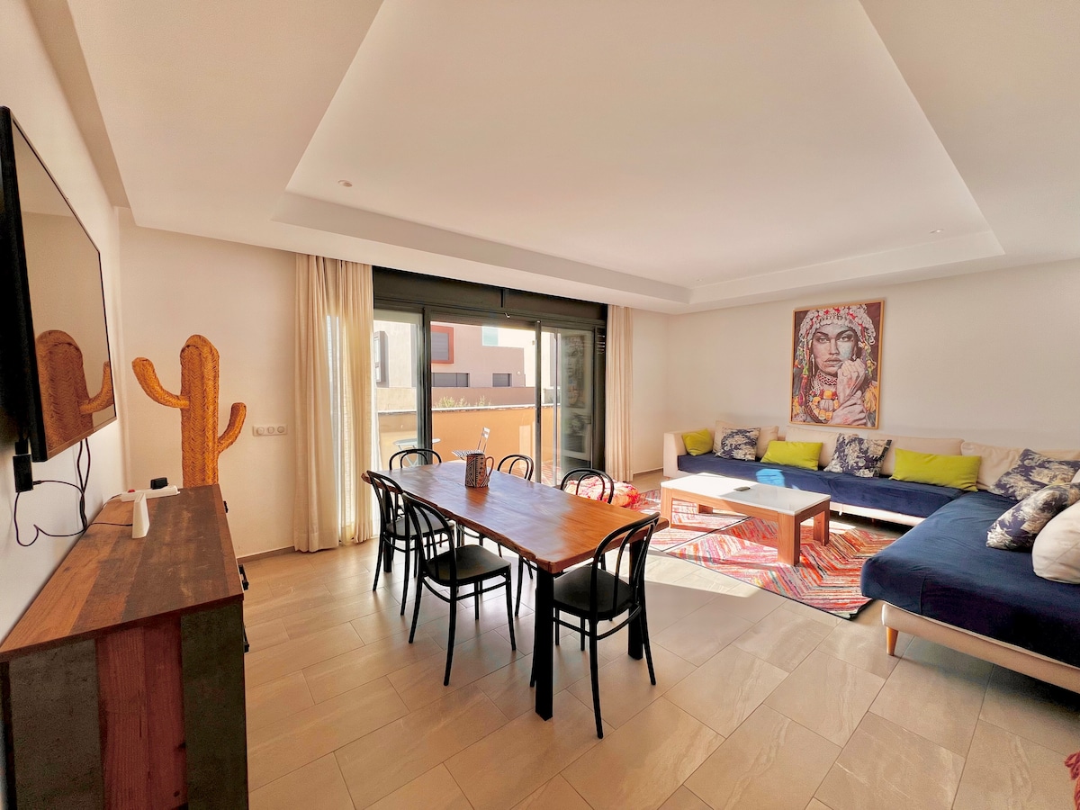 The inviting living space features a large dining table surrounded by black chairs and a comfortable seating area with colorful cushions. Large glass doors lead to a balcony, allowing natural light to fill the room. A vibrant rug adds warmth and texture to the floor.