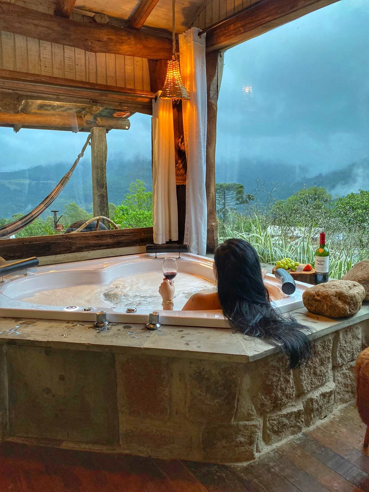 A spacious hot tub is positioned by large windows, offering views of the surrounding mountains. A relaxed individual enjoys a glass of wine, with soft lighting from a hanging lamp. Nearby, natural stone and wood accents create a tranquil atmosphere.
