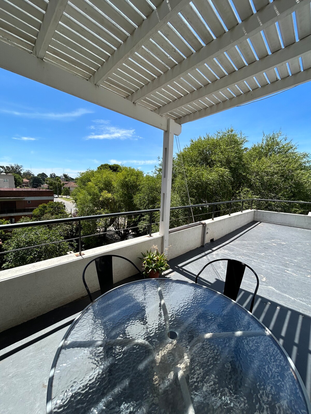 An outdoor terrace features a round glass table surrounded by black chairs. Overhead, a white pergola provides shade. Lush greenery is visible in the background, along with a clear blue sky, creating a serene space for relaxation.