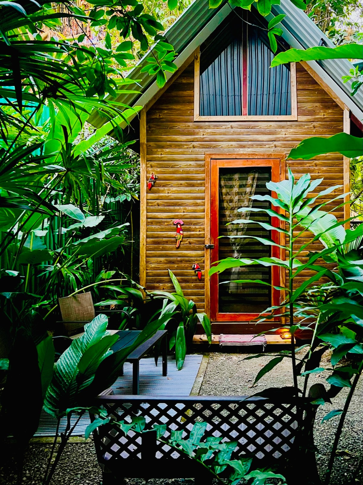 A small wooden bungalow is framed by lush tropical foliage, showcasing its natural surroundings. The entrance door features glass panels, allowing light to filter through. A seating area is visible in the foreground, inviting relaxation in this serene garden setting.