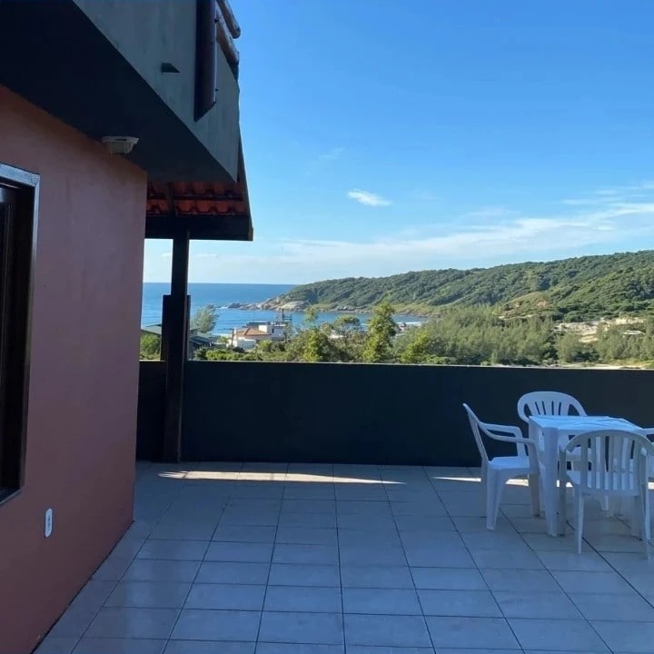 A spacious terrace is visible, featuring a table and chairs, overlooking a panoramic view of the coastline and lush green hills. The sky is clear, with a vibrant blue hue, enhancing the peaceful ambiance of the outdoor space.