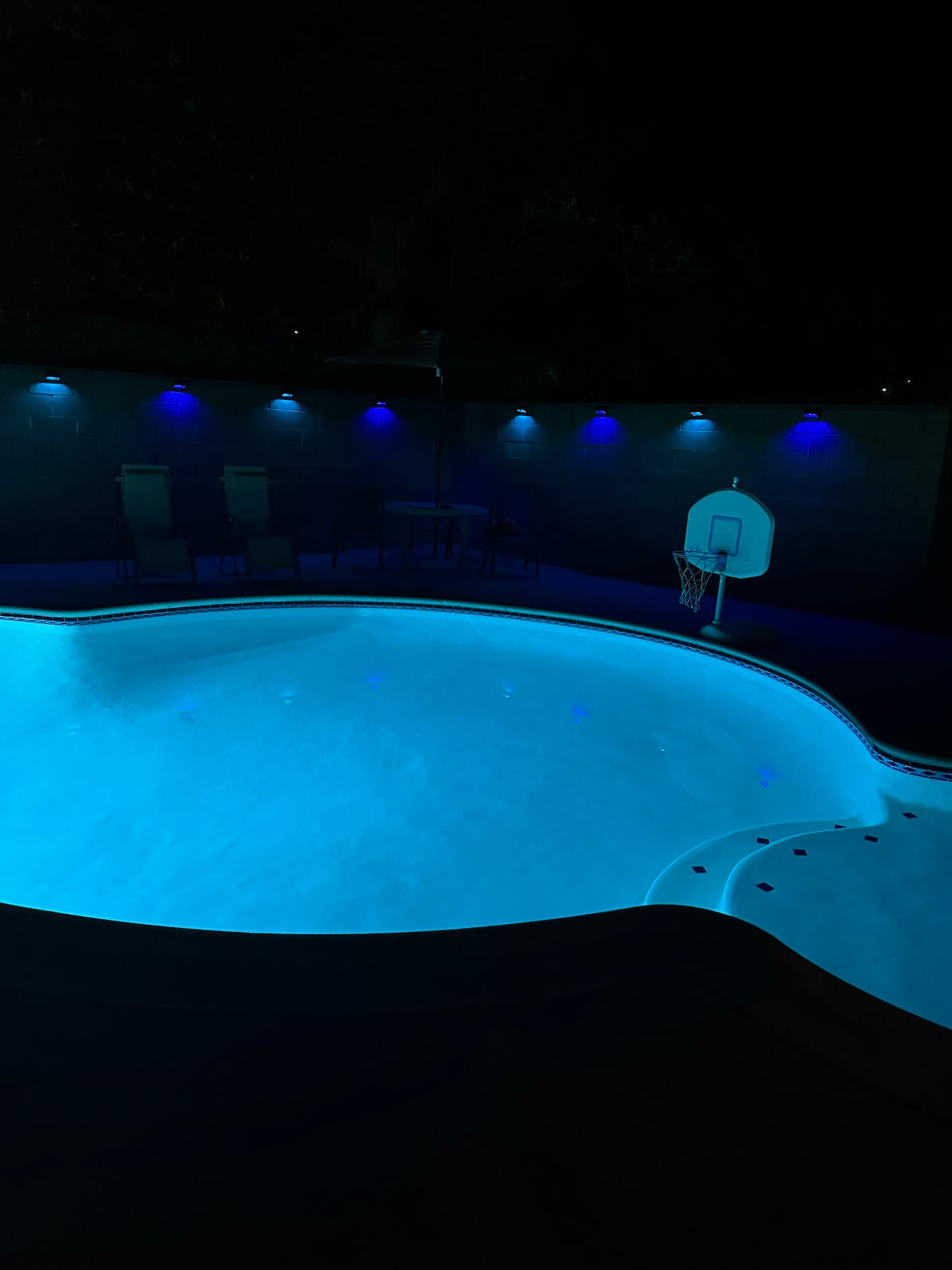 The swimming pool is illuminated in a soothing blue hue, enhancing the inviting atmosphere. A basketball hoop stands adjacent to the pool, while two lounge chairs are positioned nearby. Soft lighting casts a gentle glow around the area, creating a serene environment.
