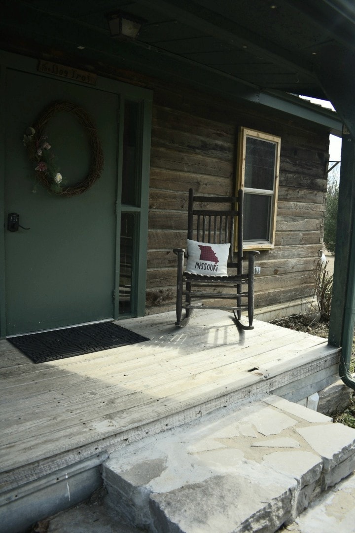 A charming wooden porch features a rocking chair with a decorative pillow. A simple wreath adorns the door, which is shaded by a roof overhang. The entrance is framed by rustic wood siding and stone steps leading up to the porch.