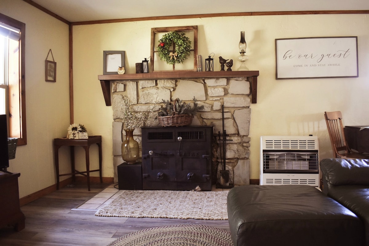 A rustic fireplace made of stone serves as a central feature in the living area. A wooden mantle displays decorative items, including a wreath and candles. A comfortable couch is positioned nearby, with a textured area rug laid on the floor, providing warmth and character.