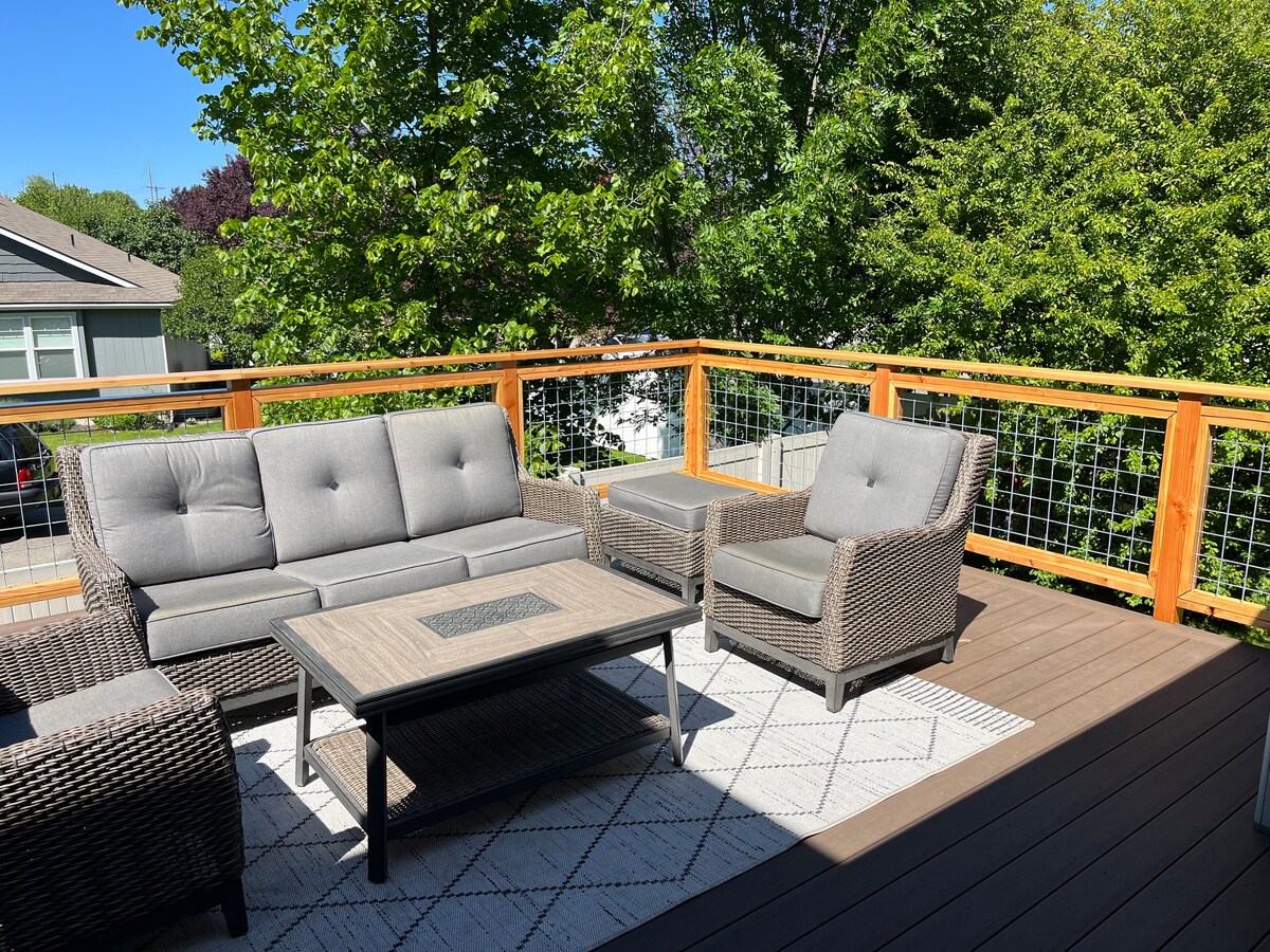 An outdoor seating area is featured on a wooden deck, furnished with a gray sectional sofa and a matching side chair. A rectangular table sits at the center, all arranged atop a decorative area rug. Lush greenery surrounds the deck, providing a serene backdrop.