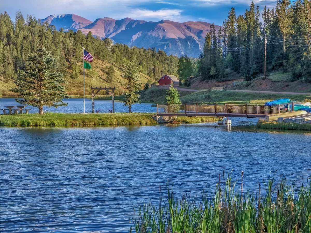 A scenic view of a tranquil lake is framed by lush greenery and tall mountain peaks in the background. The still water reflects the surrounding landscape, while a wooden dock extends into the lake, featuring a gazebo and benches. Notable elements include two flags gently waving in the breeze.