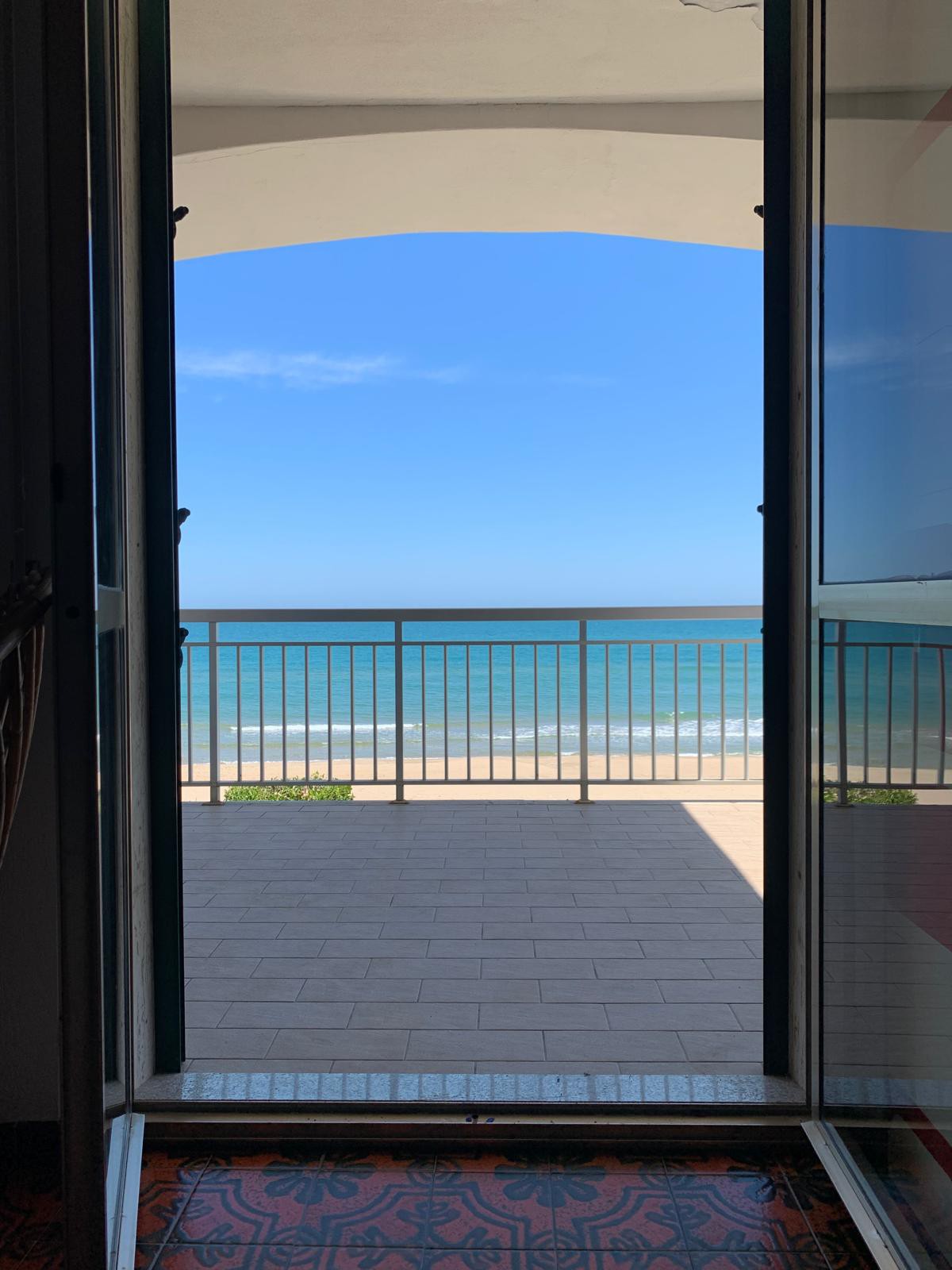 A doorway opens to a scenic view of the ocean, revealing a spacious balcony with a railing. The calm waters extend to the horizon under a clear blue sky. Natural light filters in, casting a soft shadow on the patterned floor.