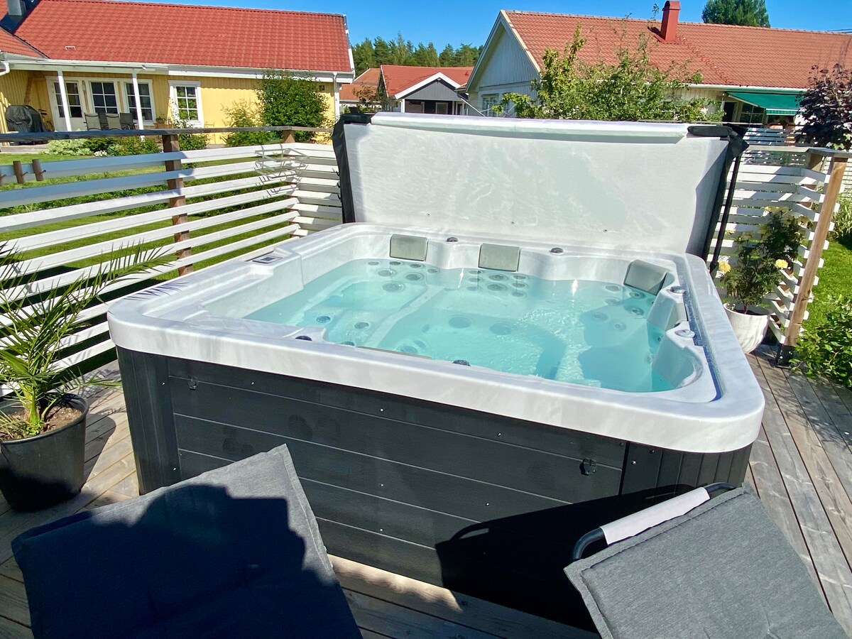A outdoor spa for six is situated on a wooden deck, showcasing a clean and inviting design. Surrounding the spa, two lounge chairs rest against a backdrop of greenery and nearby residential structures.
