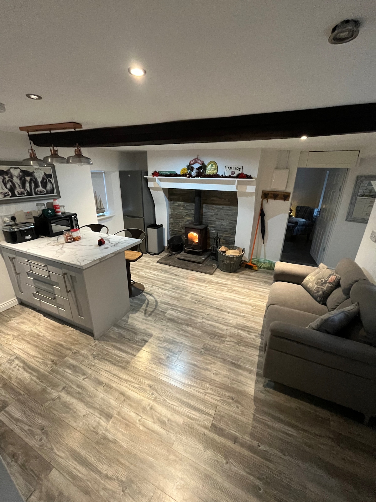 A light-filled kitchen and living area is displayed, featuring a modern kitchen island with seating and a cozy sofa. A stone fireplace is centered in the room, providing warmth. Wood-like flooring extends throughout, creating a harmonious blend of style and comfort.