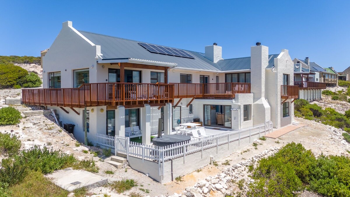 This modern two-story residence is bathed in natural light, showcasing spacious wooden decks on the upper level. Large glass doors open to the outdoor area, with a patio that includes seating. The surrounding landscape features greenery and rocky terrain characteristic of the coastal environment.