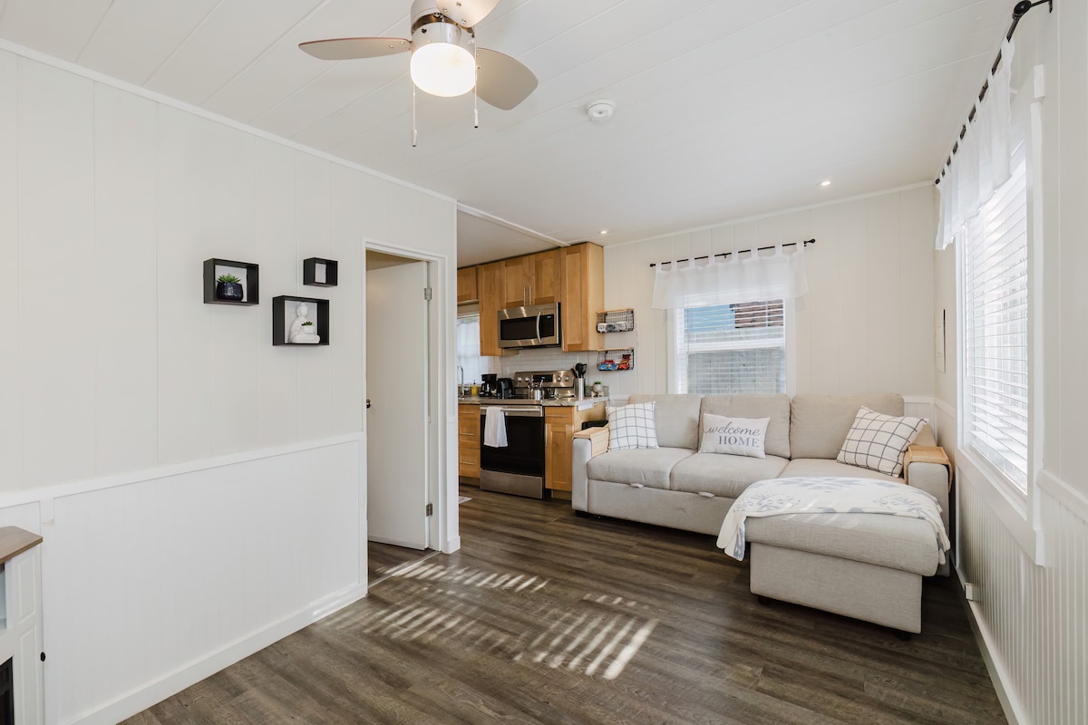 A light-filled living area is presented with hardwood floors and white walls. A sectional couch, featuring a fold-out bed, is positioned near a window dressed with sheer curtains. The adjacent kitchen includes modern stainless appliances, providing a cohesive and functional space.