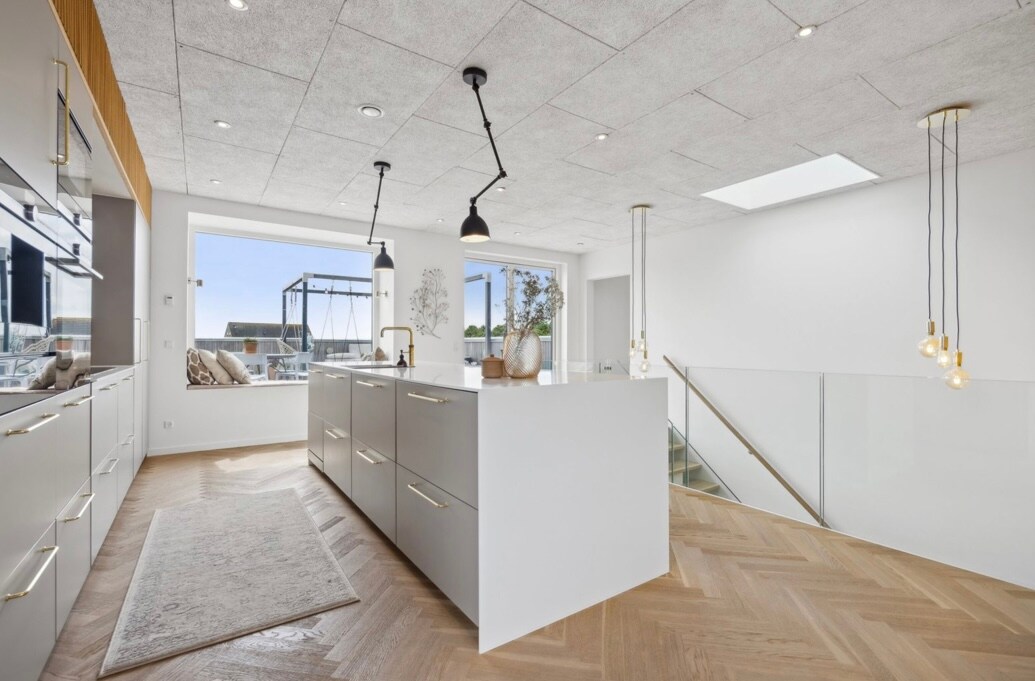 A modern kitchen space is highlighted, featuring a central island with a sleek design and ample storage. Natural light enters through the large windows, illuminating the light-colored cabinetry and herringbone-patterned wooden flooring. Decorative pendant lights hang above, enhancing the contemporary feel.