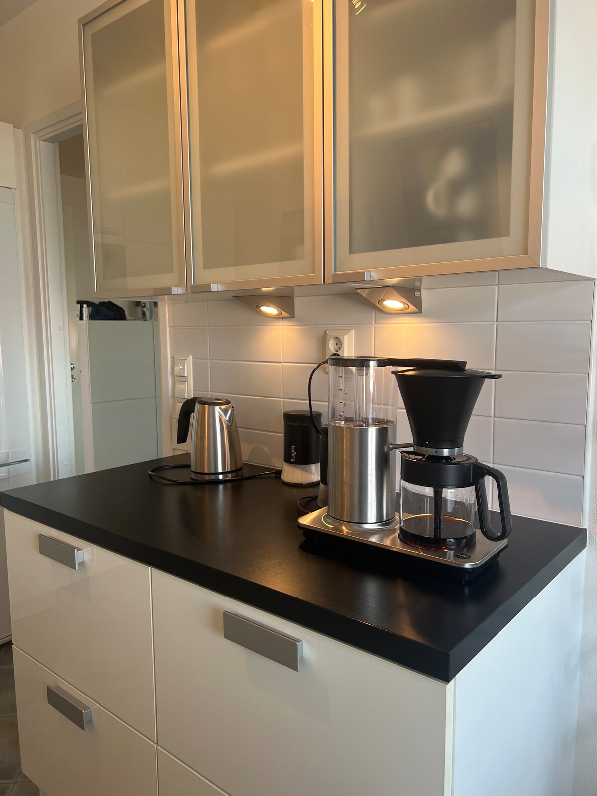 A well-equipped kitchen counter displays a kettle, a coffee maker, and a blender, all arranged on a black surface. Modern cabinetry is installed above, with glass-fronted doors showcasing items inside. Under-cabinet lights provide additional illumination.