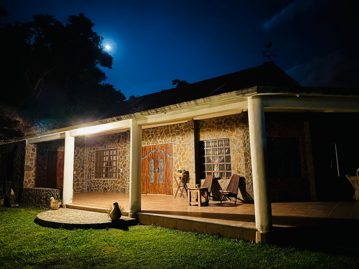 The exterior of the stone house is illuminated by soft lighting, revealing a spacious porch with wooden chairs. The moonlight enhances the serene atmosphere, while the lush grass and surrounding trees create a natural backdrop.