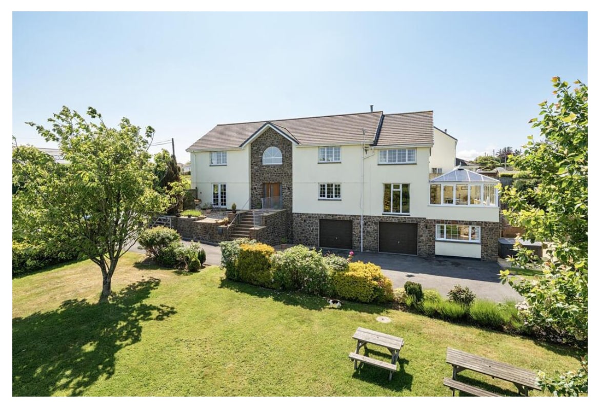 A large family house is situated amidst a well-maintained garden, featuring a mix of shrubs and grassy areas. The property showcases multiple exterior levels, a spacious driveway, and a charming conservatory that adds to the inviting appearance.