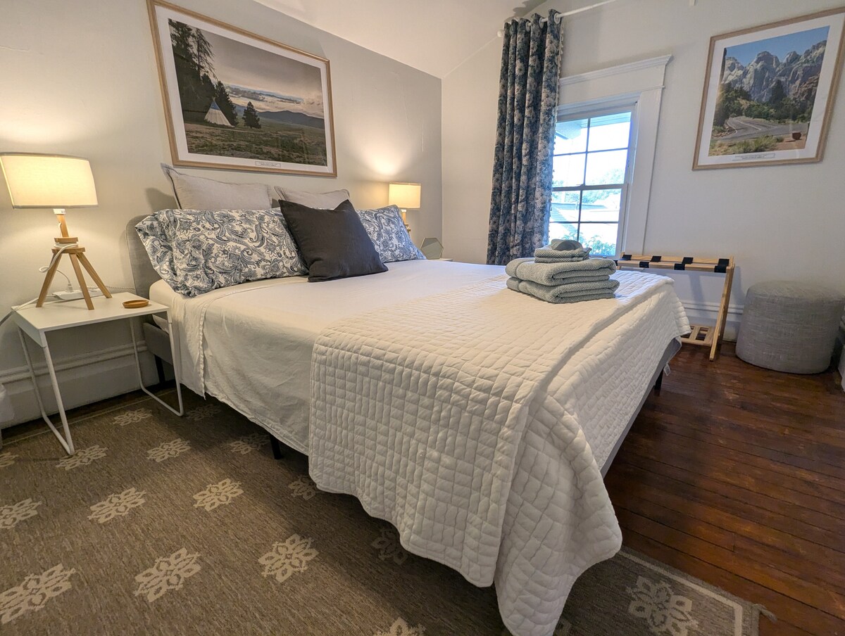 A private bedroom features a spacious bed covered with a light quilt and accent pillows. Two bedside lamps provide soft lighting. Neatly folded towels are placed on the bed. A window offers natural light, while decorative curtains frame the view. Artwork adorns the walls.