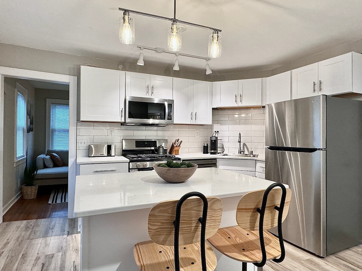 A modern kitchen features sleek white cabinetry and stainless-steel appliances. An island with two wooden stools offers additional seating. Soft pendant lighting illuminates the space, while a window allows natural light to highlight the countertop arrangement.