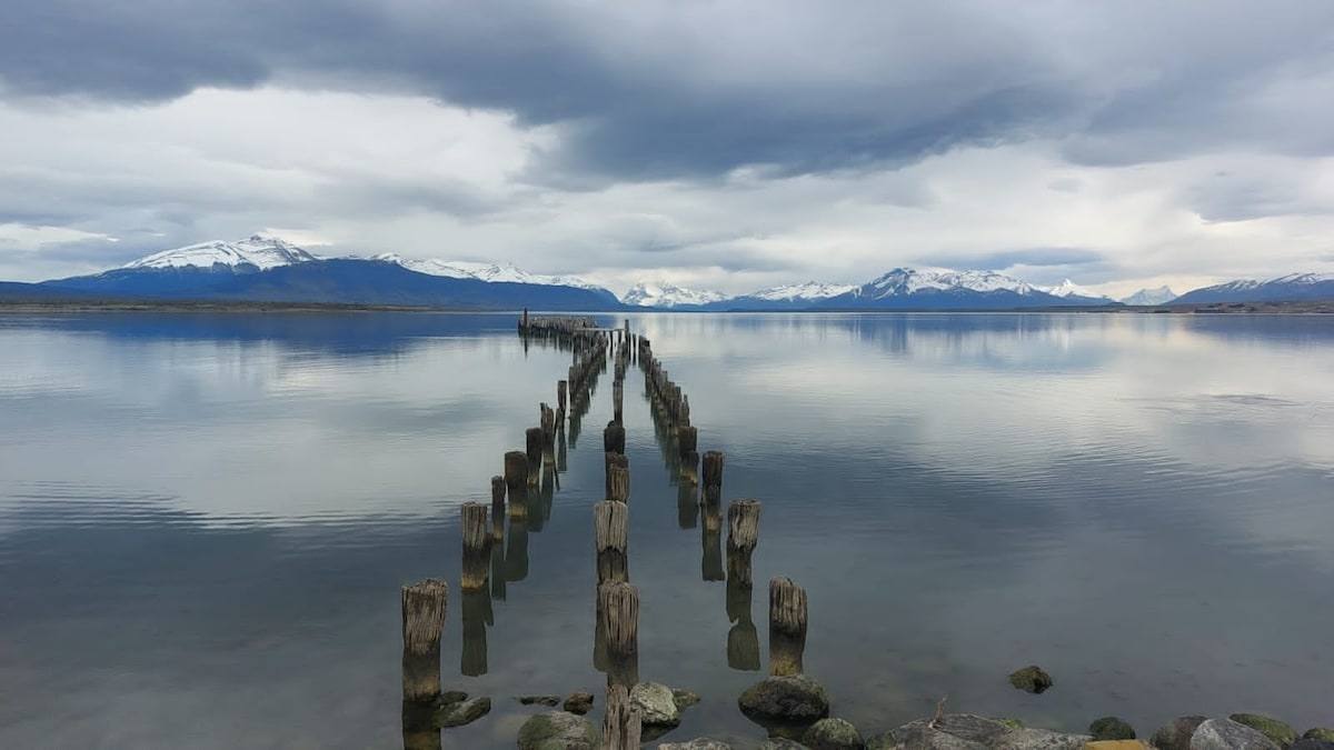A weathered pier extends into calm waters, bordered by smooth stones. The scene is framed by mountains under a cloudy sky, reflecting gently on the water's surface. The overall ambiance conveys a serene and tranquil environment.