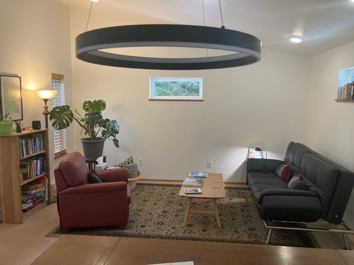 The living area features a modern design with a dark circular light fixture hanging above a wooden table. A comfortable red armchair and a sleek black sofa are positioned on a patterned rug, complemented by two small windows allowing natural light to fill the room.