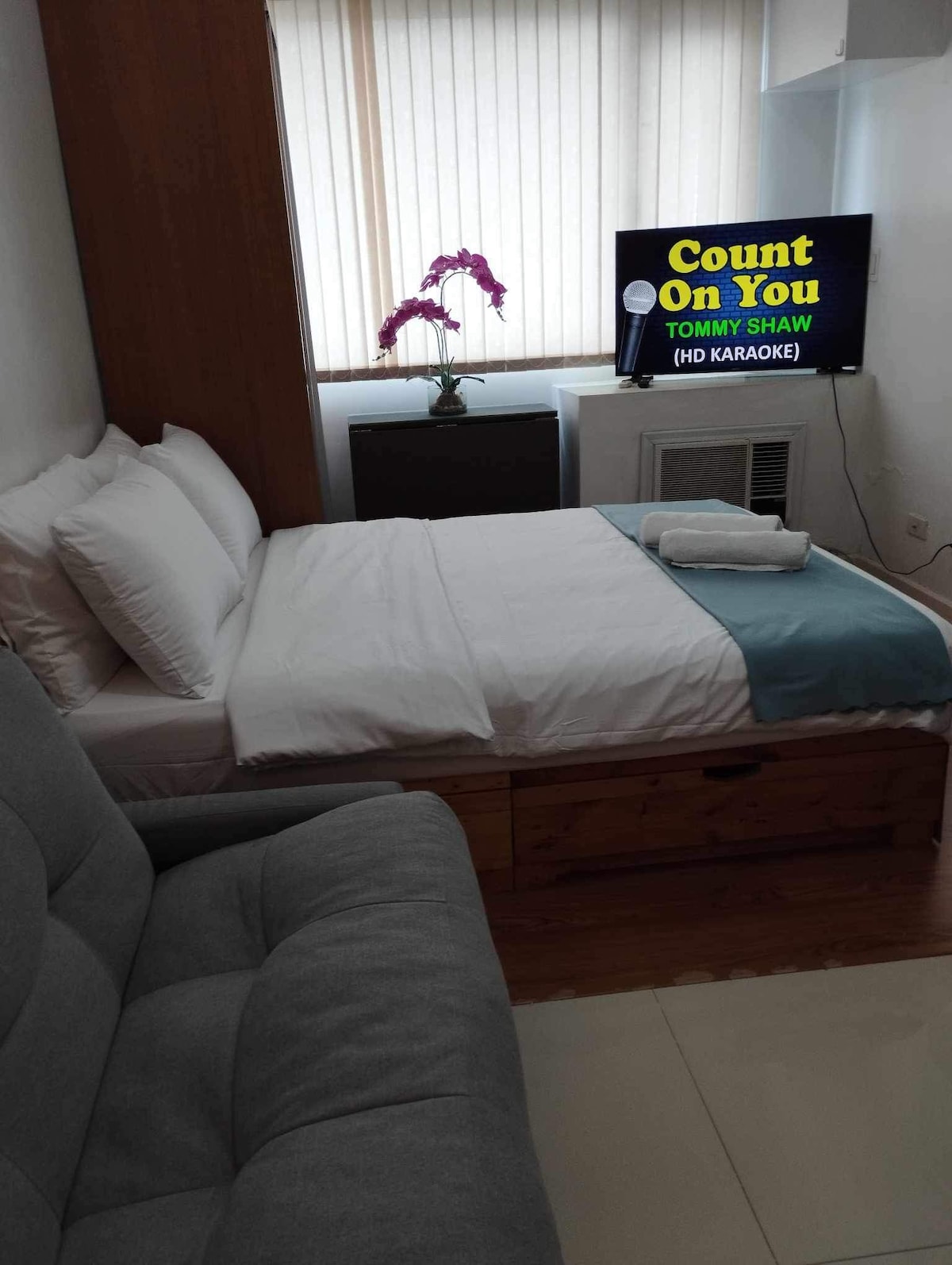 A compact bedroom features a cozy double bed with white linens and a blue throw blanket. A grey sofa is positioned nearby. A 43" smart TV is mounted on the wall, displaying a karaoke program. Natural light filters through partially closed blinds.