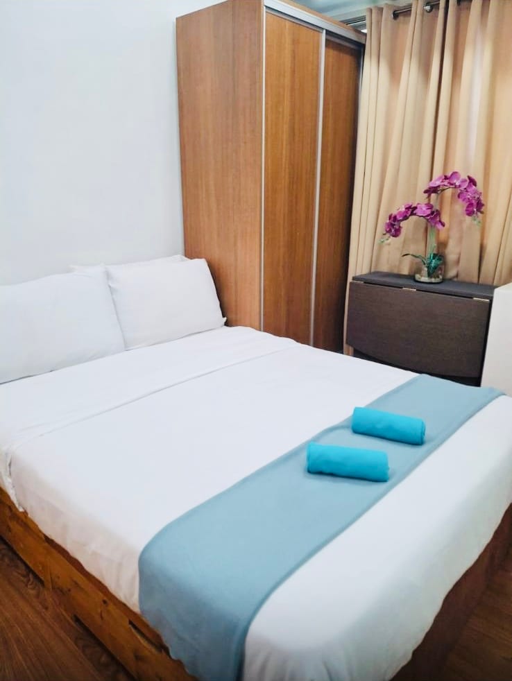 A cozy sleeping area features a double-sized bed with crisp white linens and two blue towels artfully arranged. Soft curtains in warm tones frame the space, while a wooden wardrobe and a chest of drawers add functional storage options.