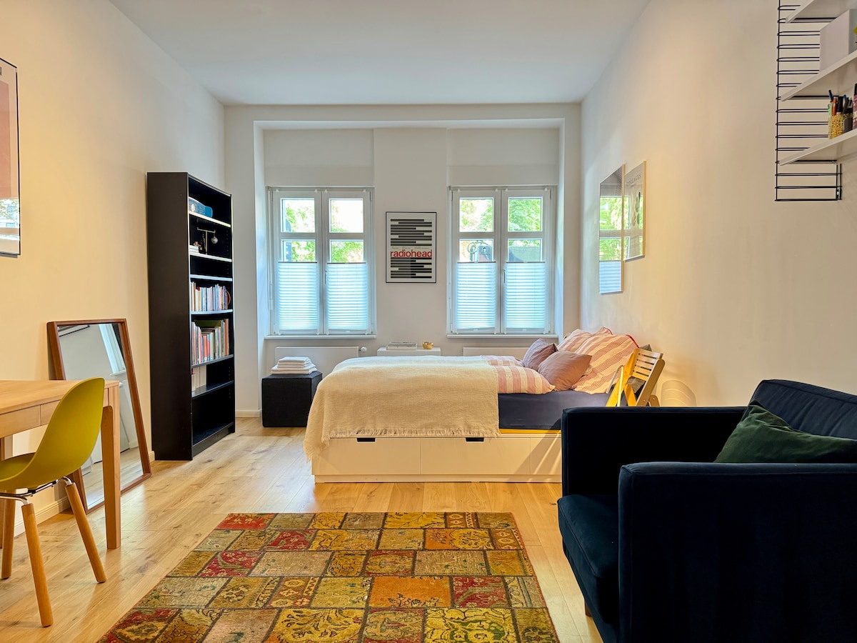 A spacious room features large windows allowing natural light to fill the space. A comfy bed with colorful bedding is positioned alongside a bookshelf filled with books. A small dining table and chairs occupy one corner, while a cozy armchair adds to the inviting atmosphere.