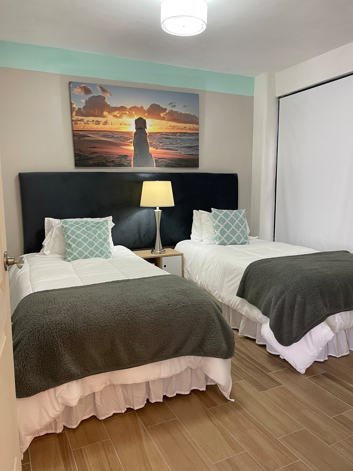 The room includes two twin beds, each dressed with white linens and dark grey throw blankets. A bedside table with a lamp is positioned between the beds. A large piece of art depicting a sunset over water is displayed on the wall, adding a serene touch.