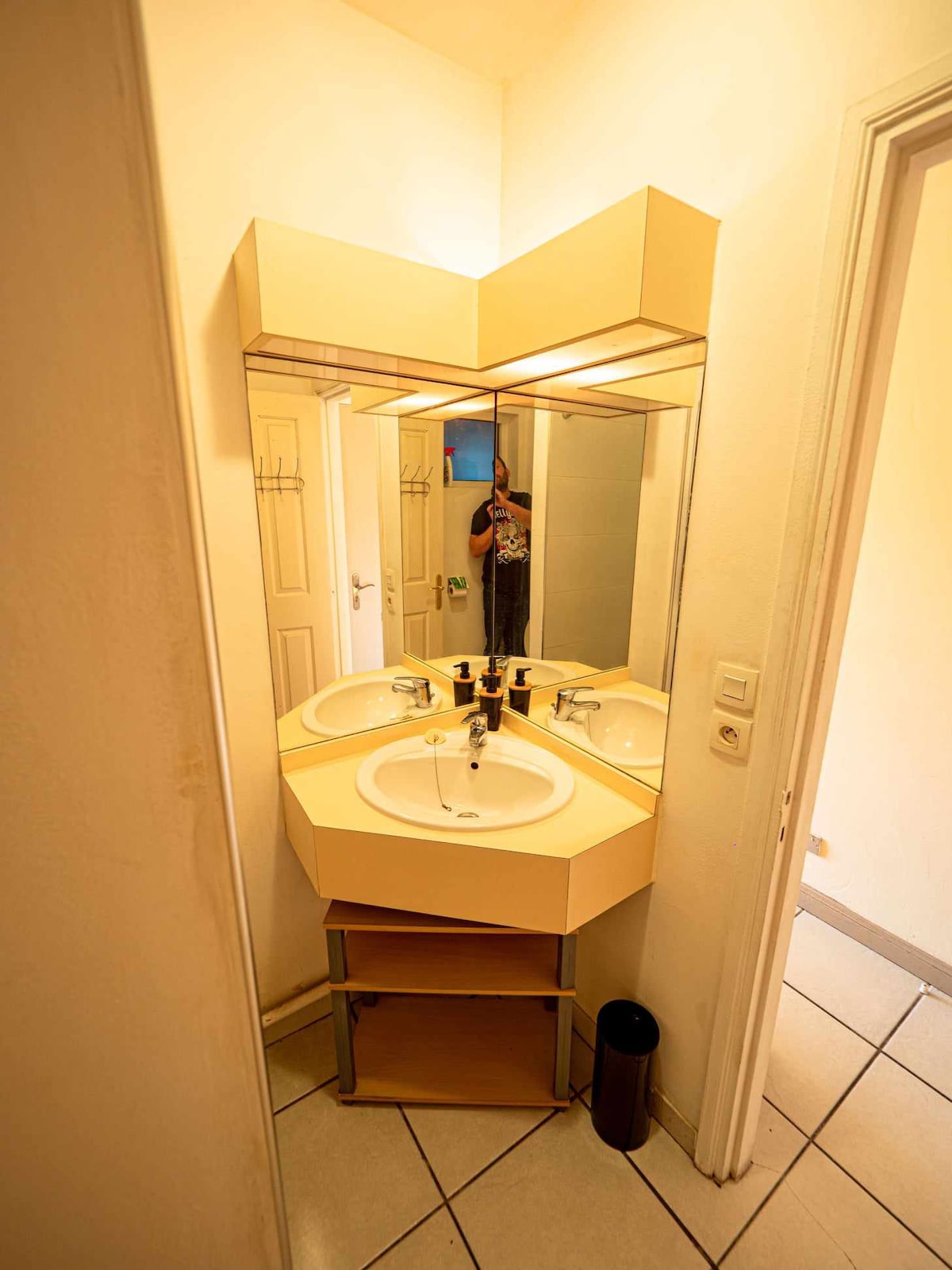 A compact bathroom area features a double sink with mirrors positioned above. The sinks are set in a hexagon-shaped countertop, and the space is well-lit, emphasizing the tiled floor and light-colored walls. A waste bin is located beside the sink setup.