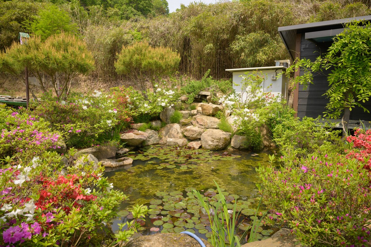 A serene garden pond is surrounded by lush greenery, vibrant flowers in a range of colors, and natural stones. Water lilies float peacefully on the water's surface, while small trees and shrubs provide a tranquil backdrop, enhancing the peaceful ambiance of the outdoor space.