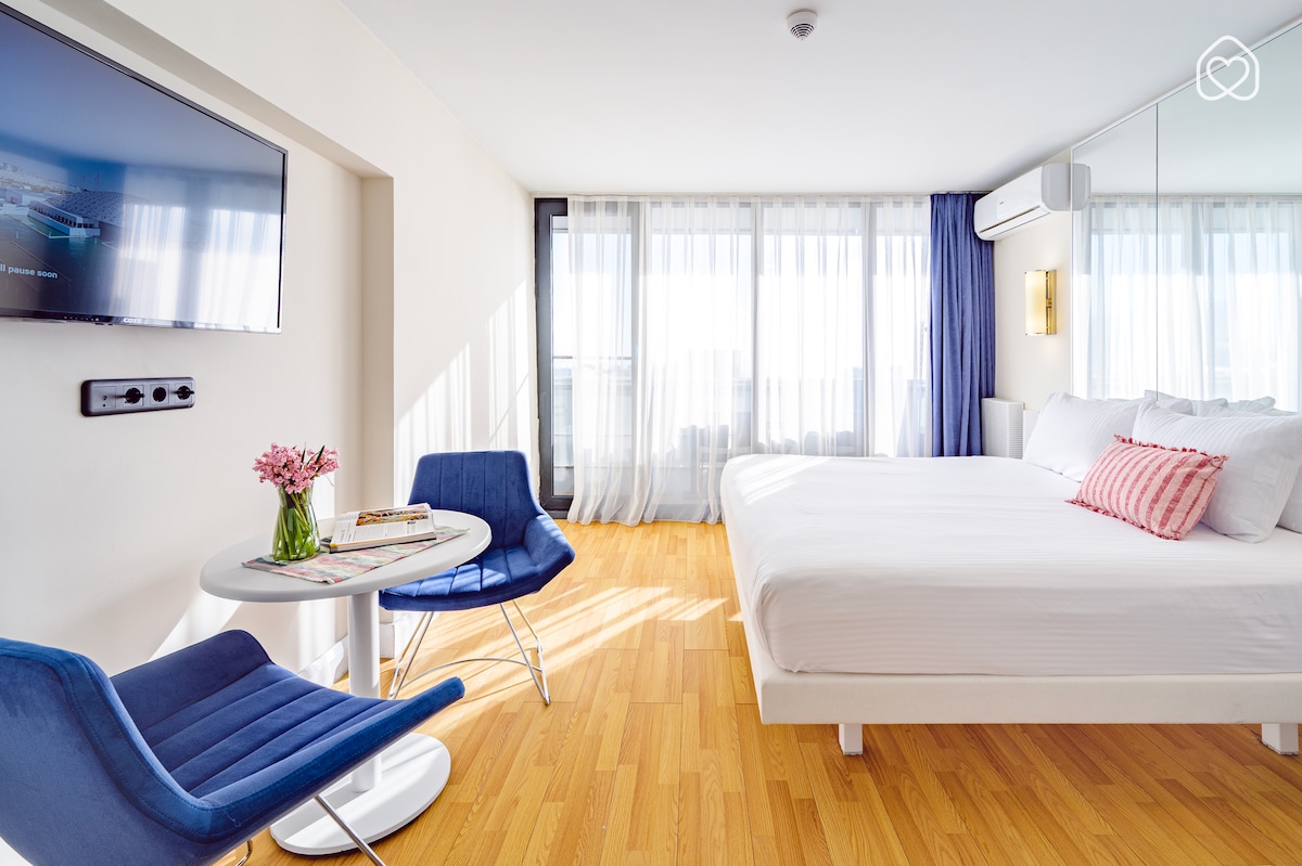 The spacious studio features a large bed with a soft white duvet and decorative pillows. Two blue chairs are positioned around a round table, while a wall-mounted TV is visible. Natural light fills the room through large windows dressed in sheer curtains.