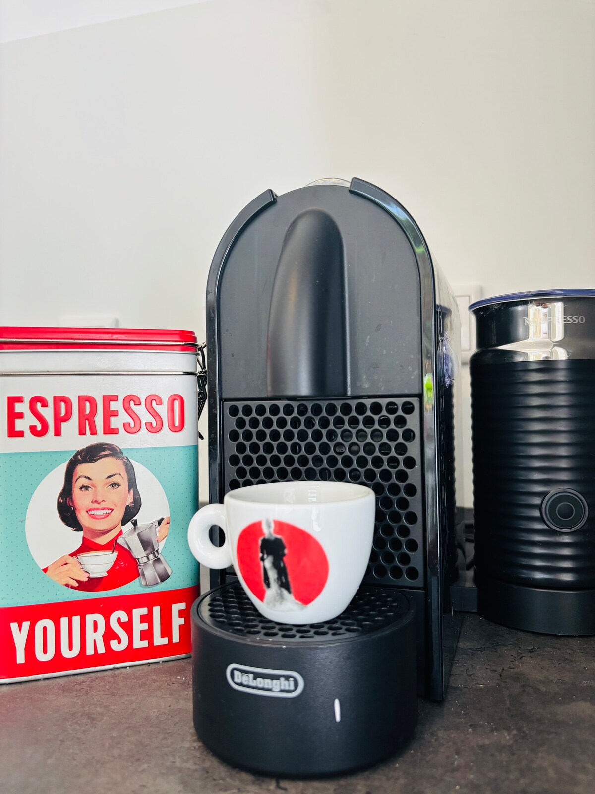 A coffee station is featured, showcasing a sleek black espresso machine beside a decorative tin that reads 'ESPRESSO YOURSELF.' A white mug with a red and black design sits on a black drip tray, contributing to the inviting coffee preparation area.