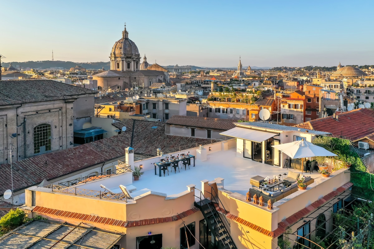 A panoramic view showcases a spacious terrace atop a penthouse, with contemporary furniture and an outdoor dining area. The stunning skyline of Rome features historic domes and rooftops, illuminated by the warm glow of sunset, creating an inviting atmosphere for evening gatherings.