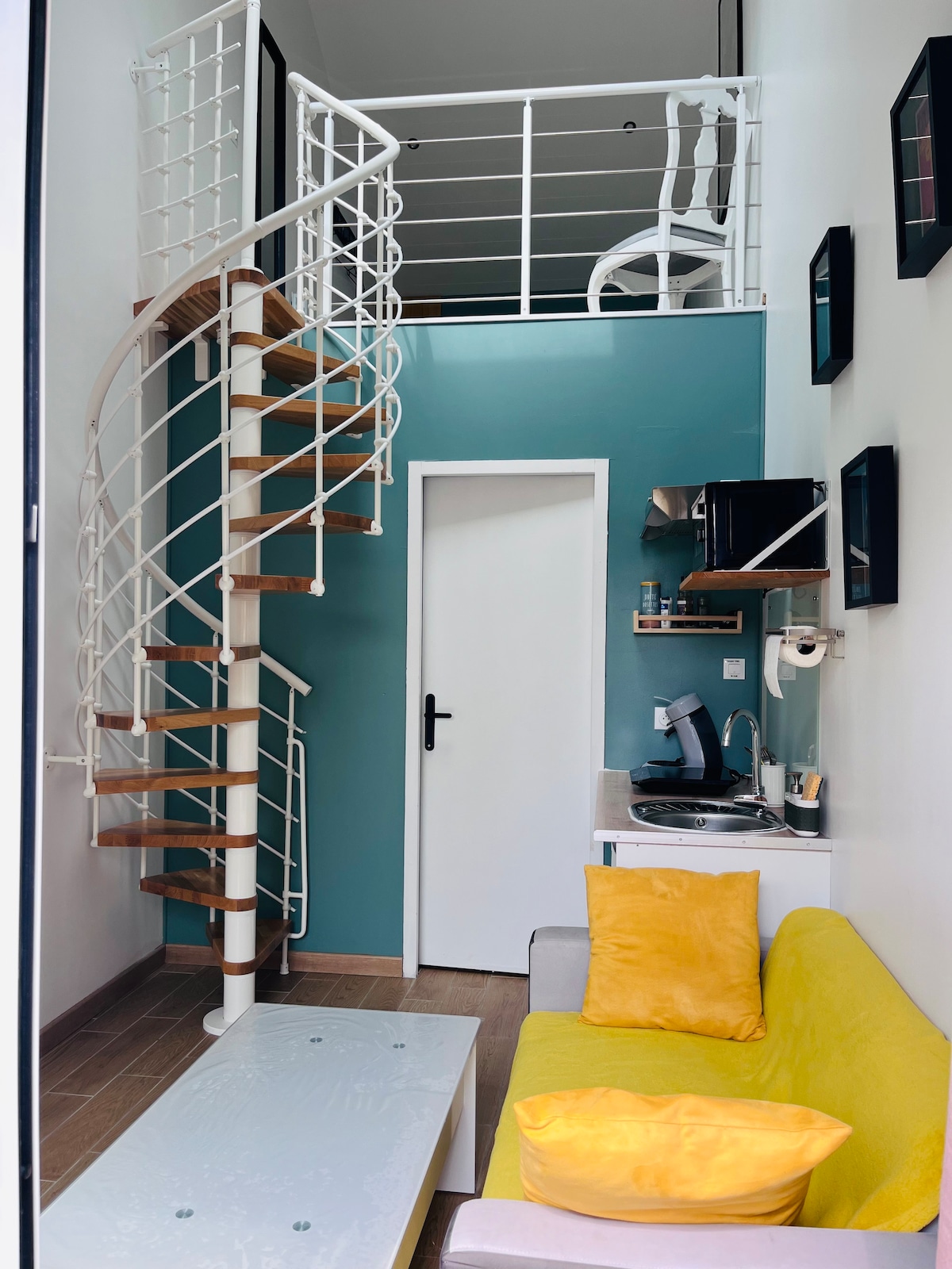 A spiral staircase leads to a vibrant upper level, enhancing the spacious feel of the home. A cozy yellow sofa is positioned in the living area near a light-colored coffee table. A compact kitchenette with appliances is visible, along with neatly arranged wall-mounted frames.