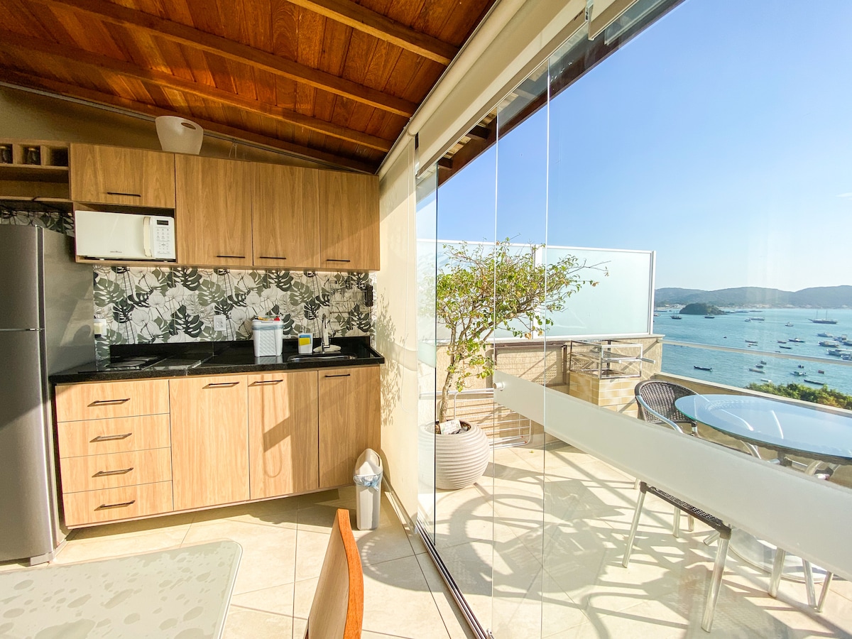 A modern kitchen area is shown with a wooden ceiling and sleek cabinets. A large window offers a stunning view of the ocean and distant boats. A dining table is positioned nearby, complementing the open, airy feel with natural light.