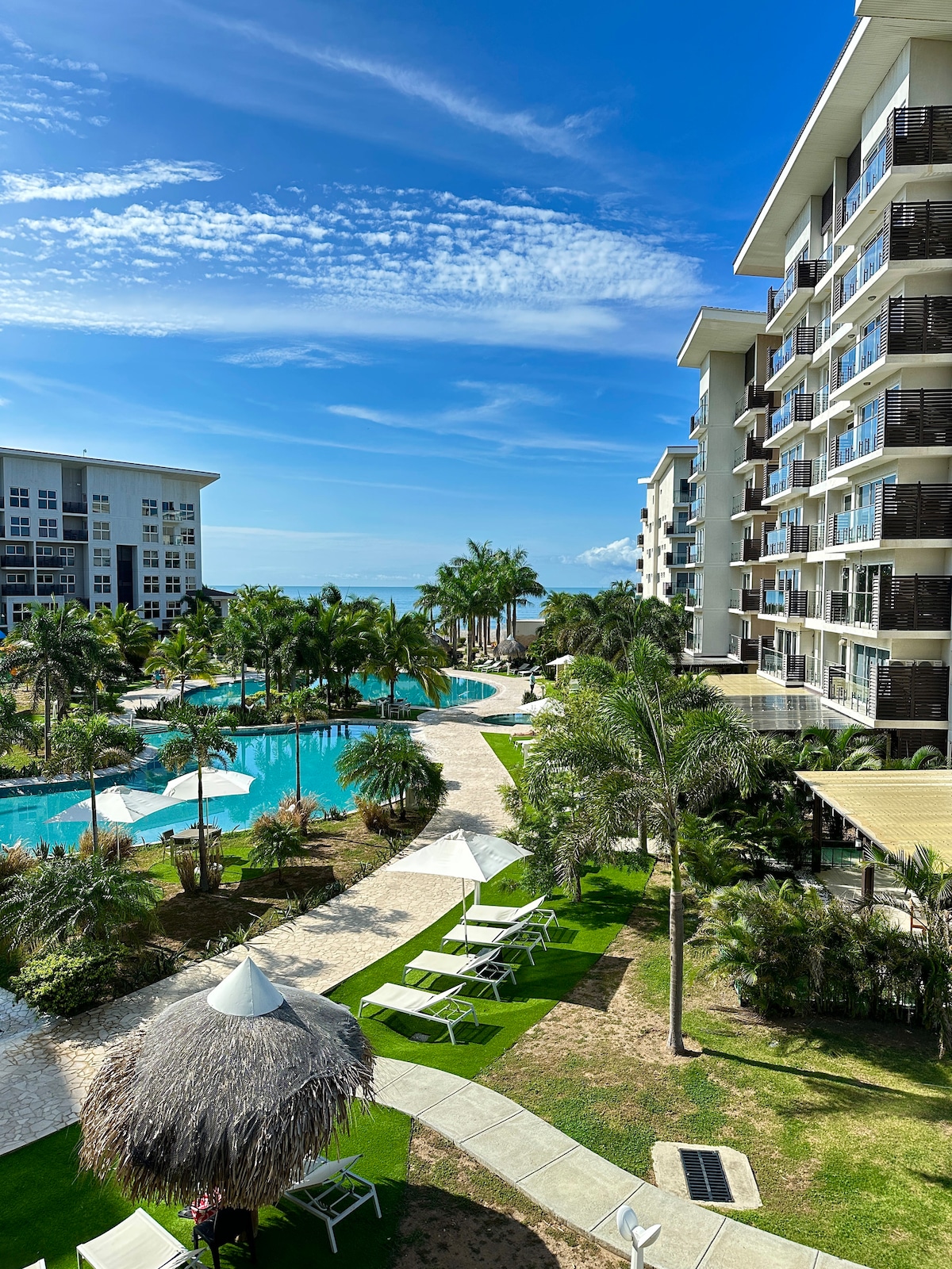 A vibrant pool area is visible, featuring palm trees and lounge chairs scattered around. A pathway winds through the lush landscaping, leading to the water. The backdrop includes modern buildings, while a clear blue sky complements the serene atmosphere.