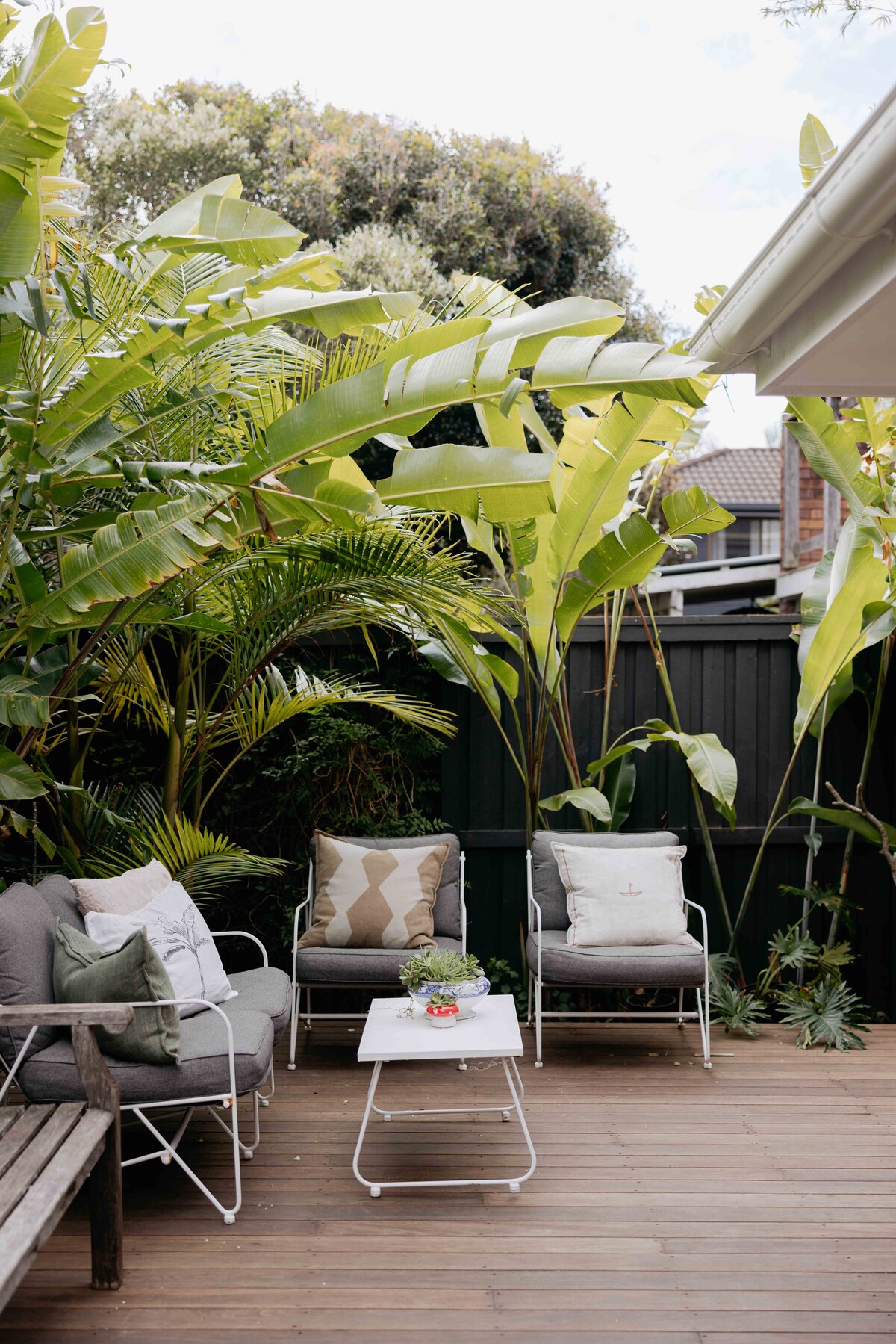 A private courtyard is surrounded by lush greenery, featuring comfortable seating with decorative cushions. A small coffee table sits at the center, creating an inviting space for relaxation amidst tropical plants.