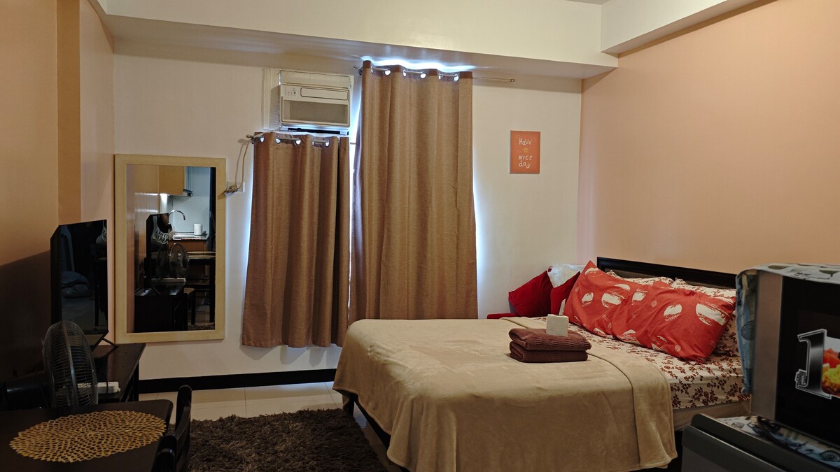 A well-appointed space features a spacious bed with decorative pillows, complemented by a light blanket. Two large curtains frame a window, allowing natural light to filter through. A flat-screen TV rests on a stand across from the bed, with a sofa and table positioned nearby.