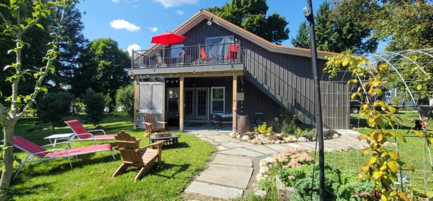 A modern barn-style exterior is surrounded by well-maintained gardens and a grassy area. A spacious deck with red umbrellas overlooks the property. Lounge chairs and a fire pit are present in the outdoor space, inviting relaxation and enjoyment of the surroundings.