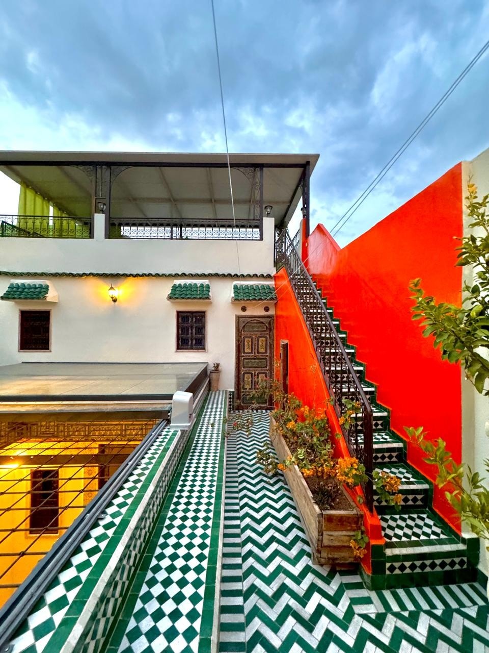 A vibrant staircase is shown leading to a rooftop terrace, flanked by charming decorative tiles in green and white. The surrounding walls feature a bold orange hue, contrasting with the tiled pathway and the architecture's intricate details, enhancing the space's character.