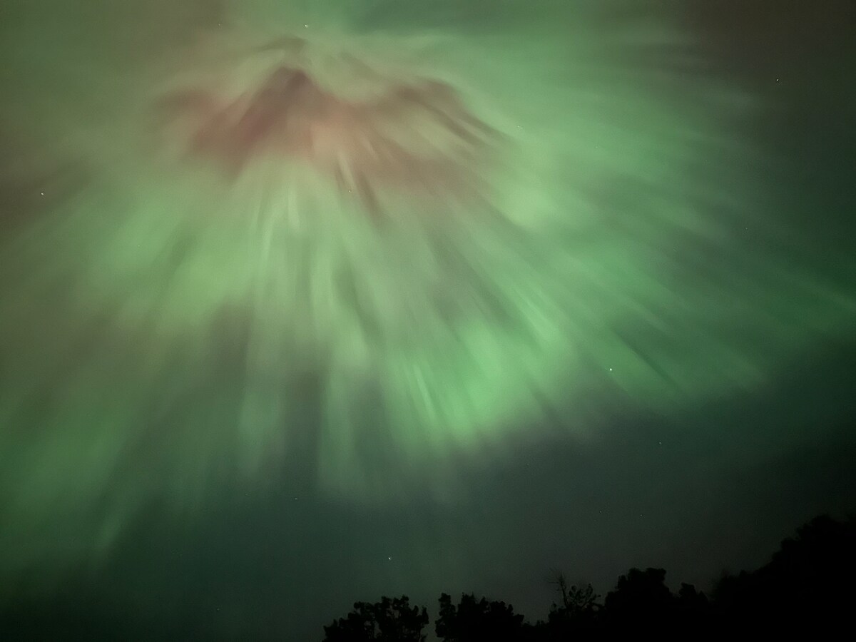 A captivating night sky is filled with swirling green and faint red hues, resembling dynamic cloud formations. The shadowy outlines of trees can be seen in the foreground, creating a contrast against the colorful celestial display above.