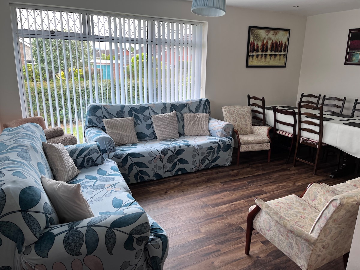 A welcoming living area features two patterned sofas complemented by a floral armchair. A wooden dining table with multiple chairs is visible nearby. Large windows allow natural light to brighten the room, enhancing the view of the greenery outside.
