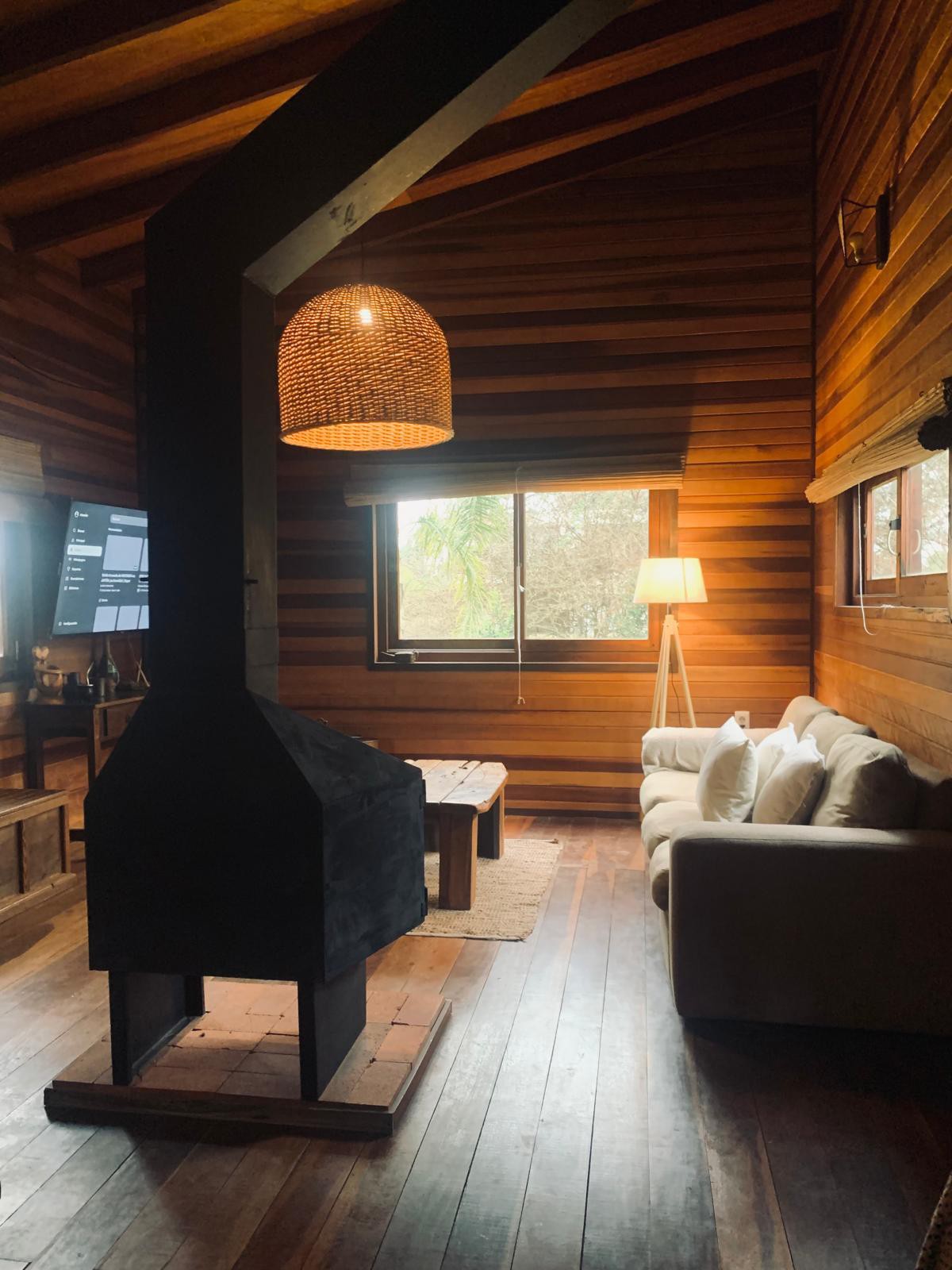 The interior of the cabin showcases wooden walls and ceilings, creating a warm atmosphere. A modern, black wood stove is positioned centrally, with a comfortable sofa nearby. Natural light is allowed in through a large window, complemented by a standing lamp.