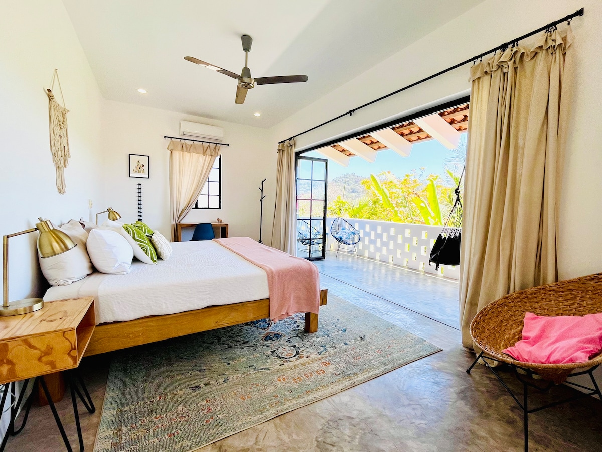 A spacious bedroom features a king bed with crisp white linens and colorful accent pillows. Large glass doors open to a terrace, allowing natural light to fill the room. A rustic rug adds warmth, while a fan provides air circulation. Light curtains frame the doorway.
