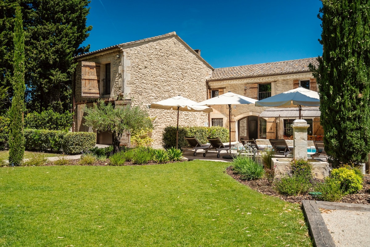 A charming residence is showcased, featuring natural stone walls and multiple levels. The landscaped garden includes sun loungers and umbrellas, surrounding a tranquil pool area. Tall green trees and shrubs enhance the outdoor space, creating a serene environment for relaxation and enjoyment.